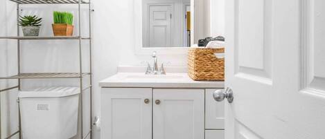 A small, white bathroom with a toilet, a sink, and a mirror. There are shelves with plants and a basket on the sink counter.
