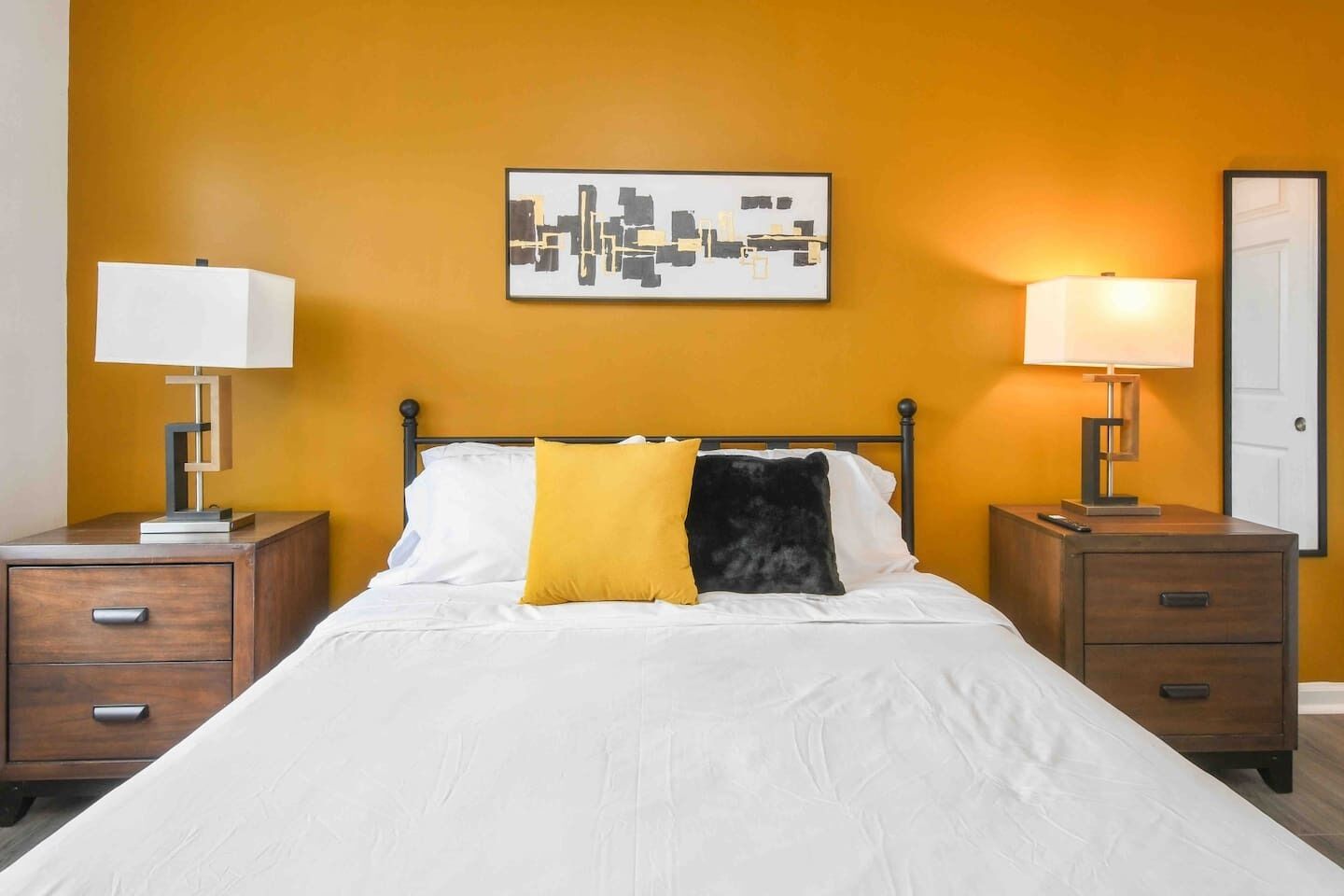 Una habitación de hotel con cama, dos mesitas de noche, una lámpara de noche, una obra de arte en la pared y una puerta.