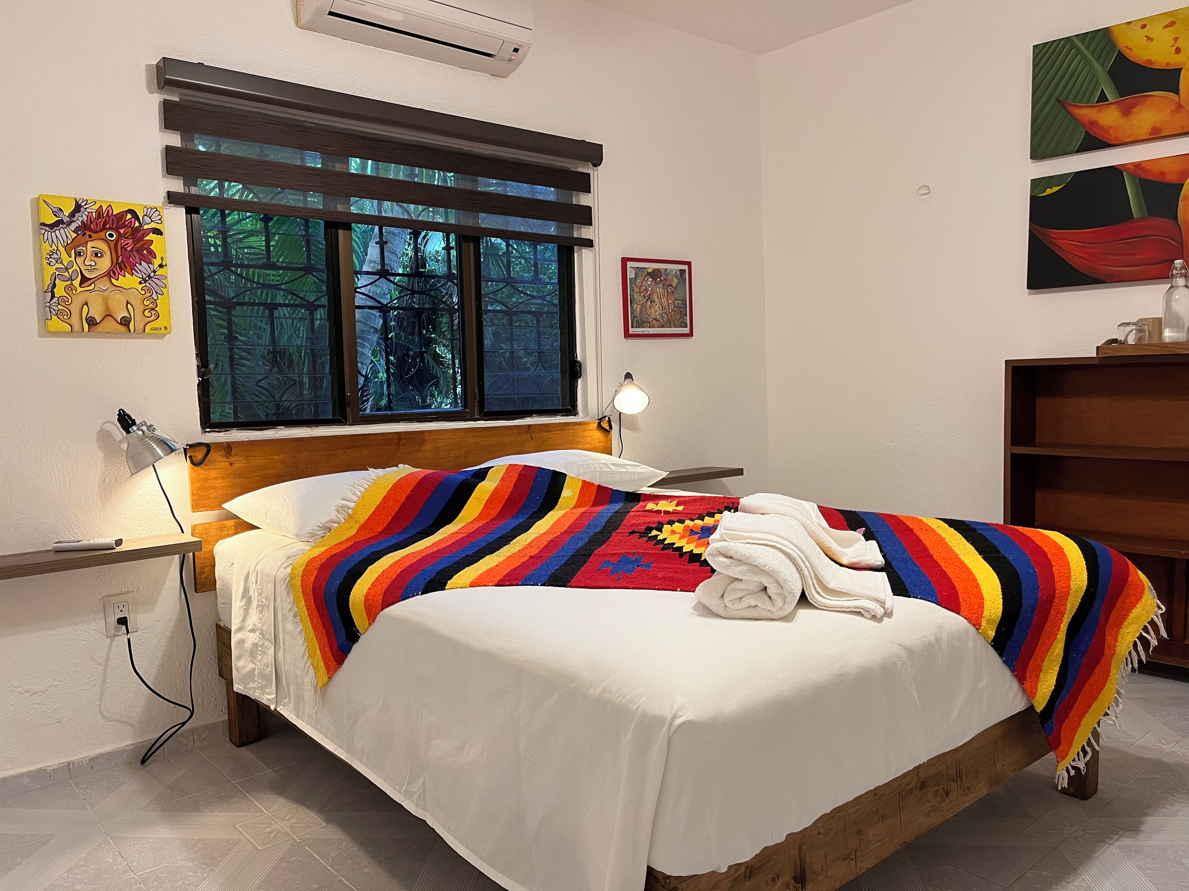 A bed with a white sheet and a colorful striped blanket, a bedside lamp, and a window with blinds.