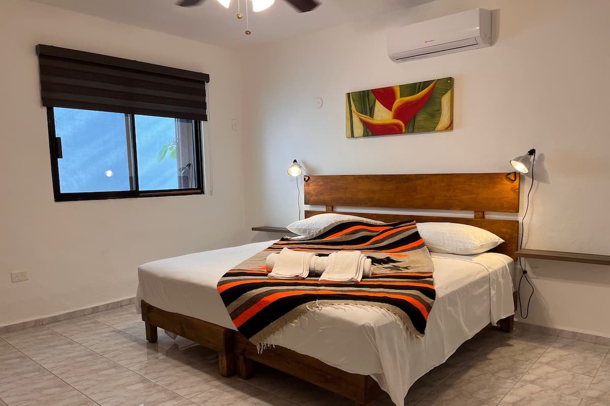 Ein Schlafzimmer mit einem hölzernen Bett, weißer Bettwäsche, einer farbenfrohen Decke, einem Kopfteil mit Wandleuchten und einem Fenster mit Jalousien sowie einer Klimaanlage.