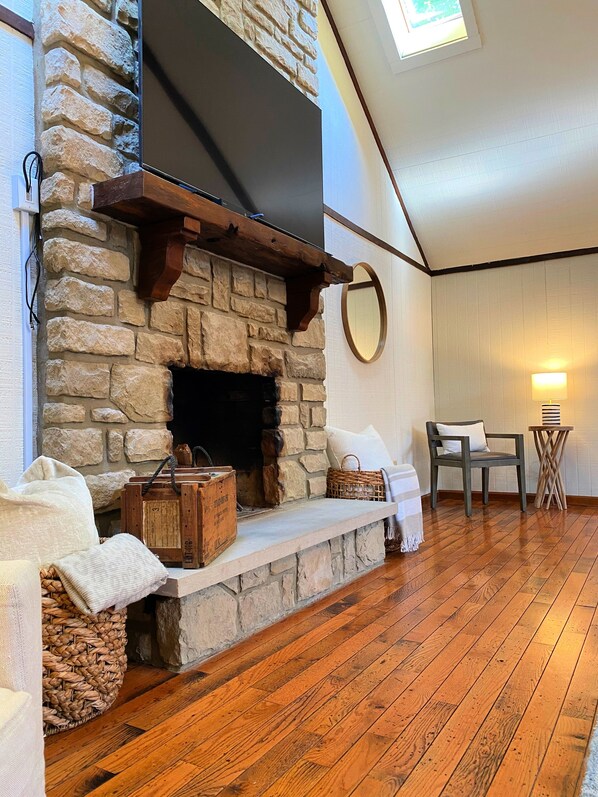A cozy room with a stone fireplace, wooden flooring, a comfortable chair, and a small table. - Hocking Hills Villa · Arcade · Hot Tub · 50 Ac · Pets (Laurelville)