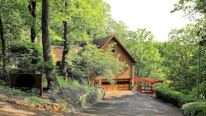 Une cabane en bois avec un garage, entourée d’une forêt dense et accessible par une allée de gravier.