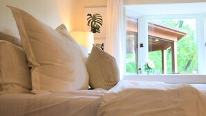 Un lit avec une literie et des oreillers blancs, une lampe de chevet et une vue sur la verdure par la fenêtre.