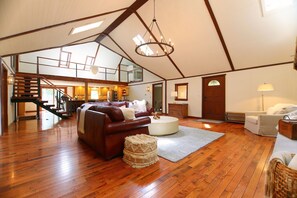 A spacious loft with a wooden floor, a brown leather sofa, a white round coffee table, and a staircase leading to an upper level. - Hocking Hills Villa · Arcade · Hot Tub · 50 Ac · Pets (Laurelville)