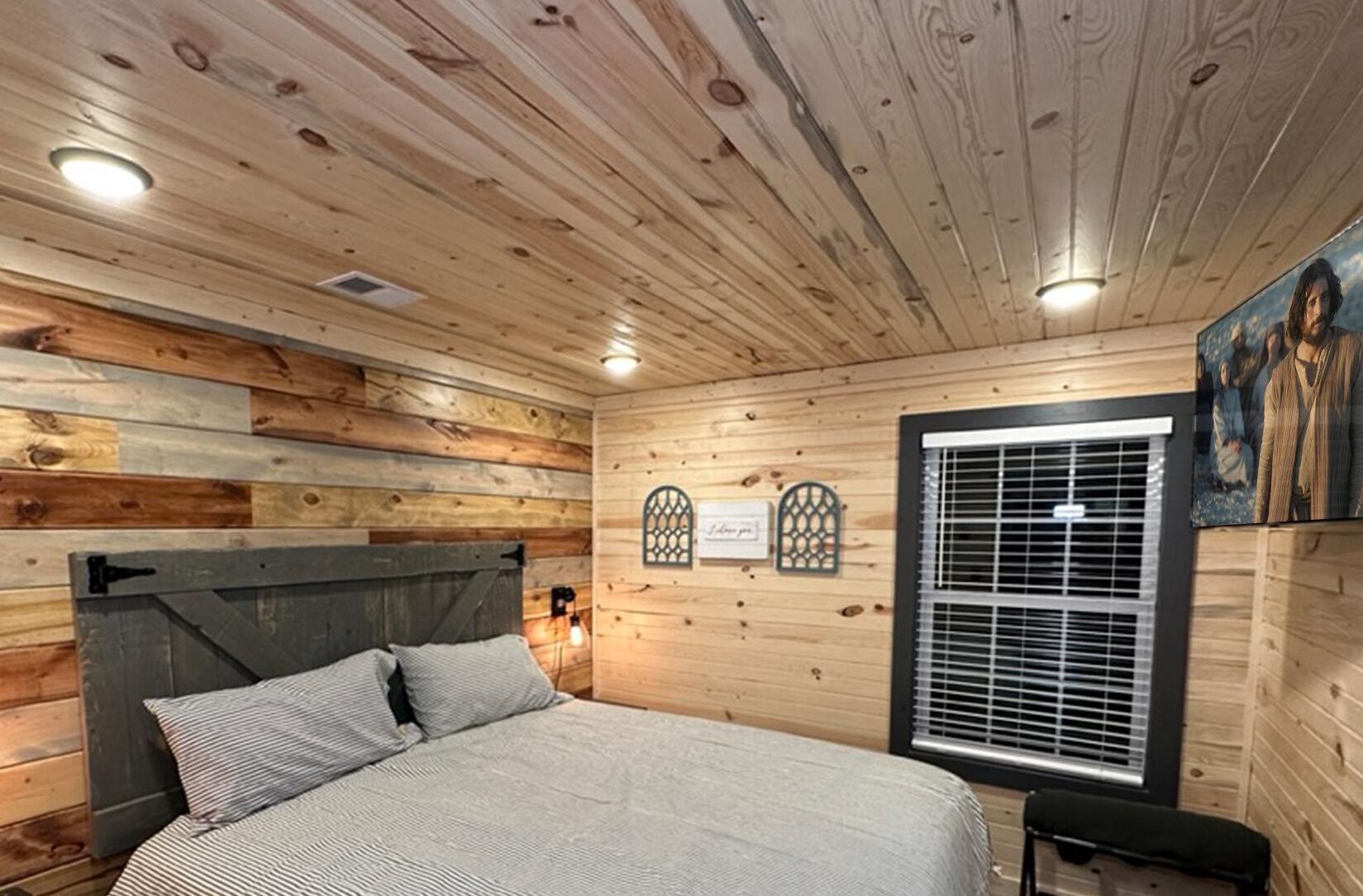 A bedroom with a wooden ceiling, a bed with striped bedding, a bench, and a window with blinds.
