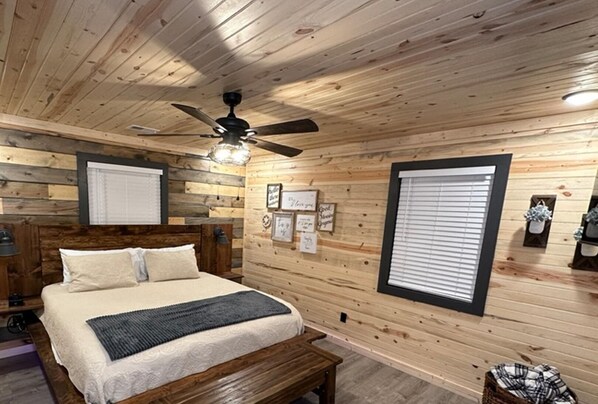 A cozy bedroom with a wooden ceiling, a bed with a headboard, a bedside table, a ceiling fan, and a window with blinds.