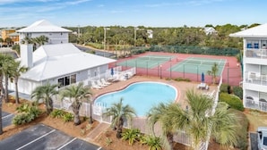 Zona de piscina con cancha de tenis, palmeras y un edificio blanco con techo inclinado.