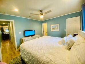 Ein Schlafzimmer mit einem Bett, einem Fernseher auf einer Kommode, einem Deckenventilator und einer Tür zu einem anderen Raum.