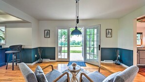 A spacious living room with a large window, a dining area, and a kitchen visible through the glass doors.
