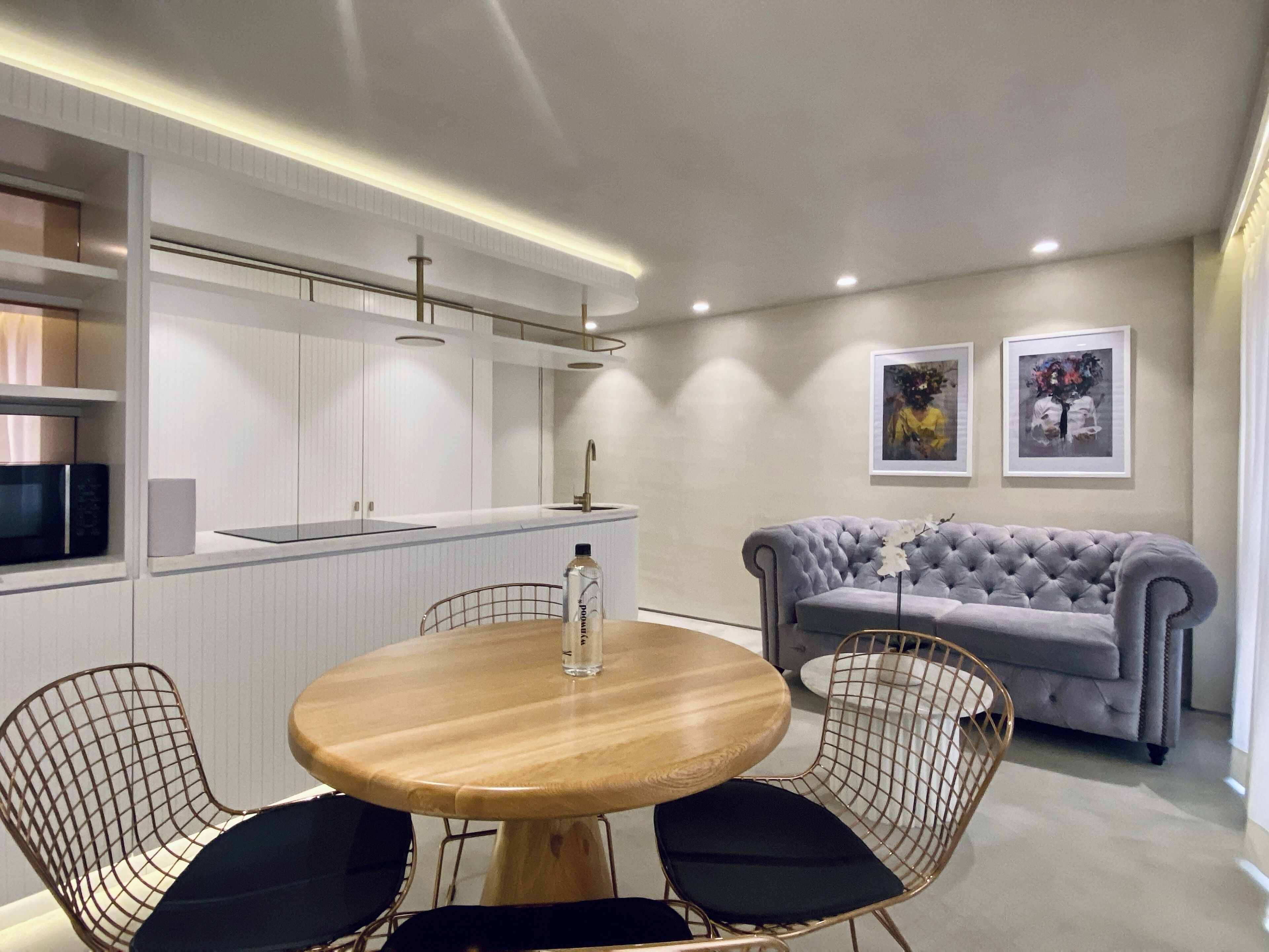 A modern kitchen with white cabinetry, a round wooden table with black chairs, a grey sofa, and two framed artworks on the wall.