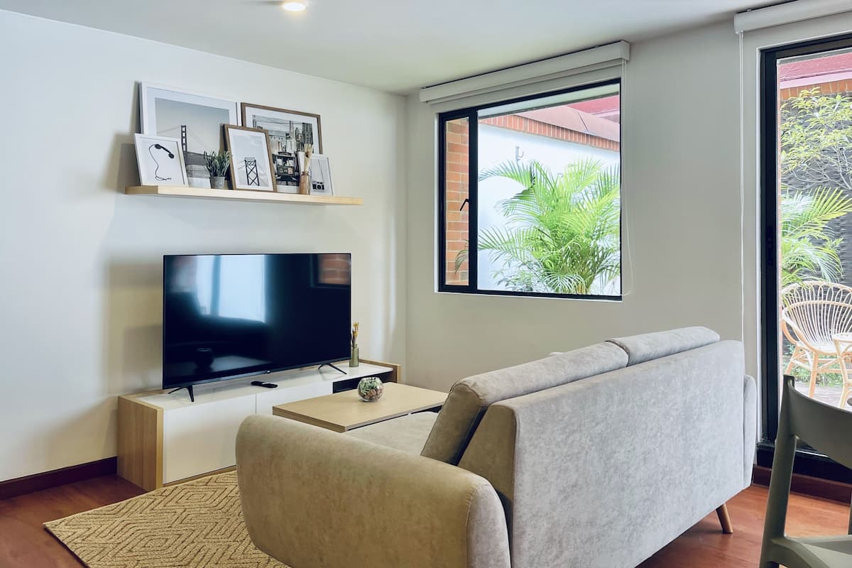 A modern living room with a flat-screen TV, a sofa, a coffee table, and a window with a view of greenery.