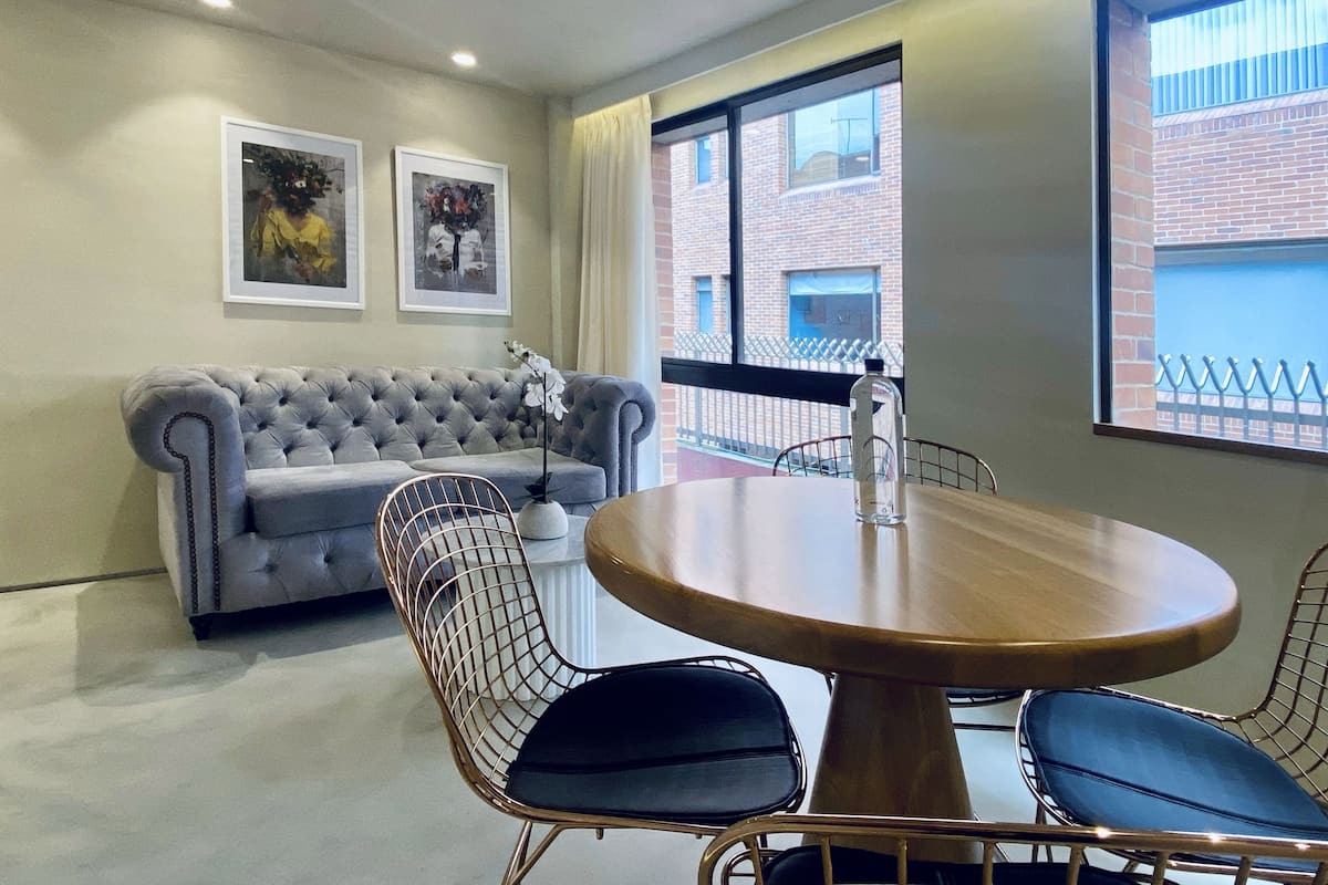 A conference room with a round wooden table, four wireframe chairs, a tufted grey sofa, and two framed artworks on the wall.