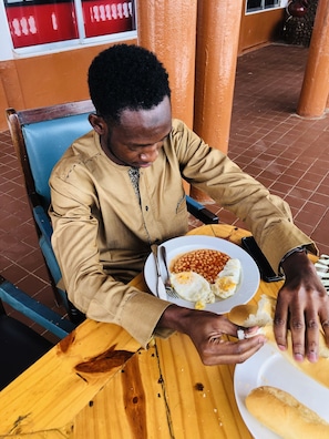 A person seated at a wooden table with a plate of food, a smartphone, and a fork. - Atlanticoast Residence (Bakau)