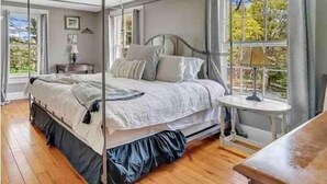 A bedroom with a four-poster bed, a bedside table with a lamp, and a window with a view of trees. - Colonial yellowhouse (Newtown)