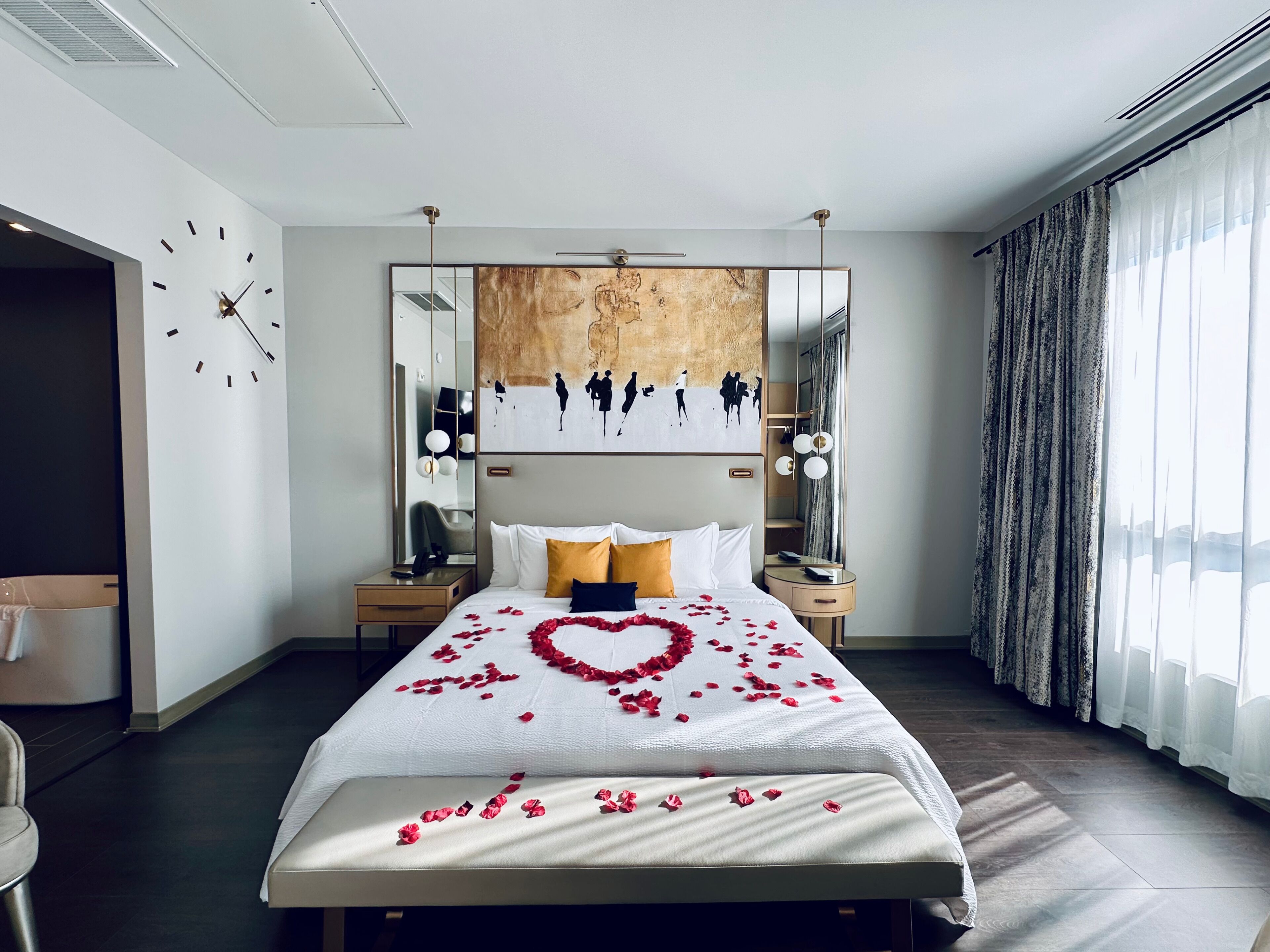 Una camera d'albergo moderna con un letto grande, una sveglia al muro e vista sulla città attraverso tende leggere.