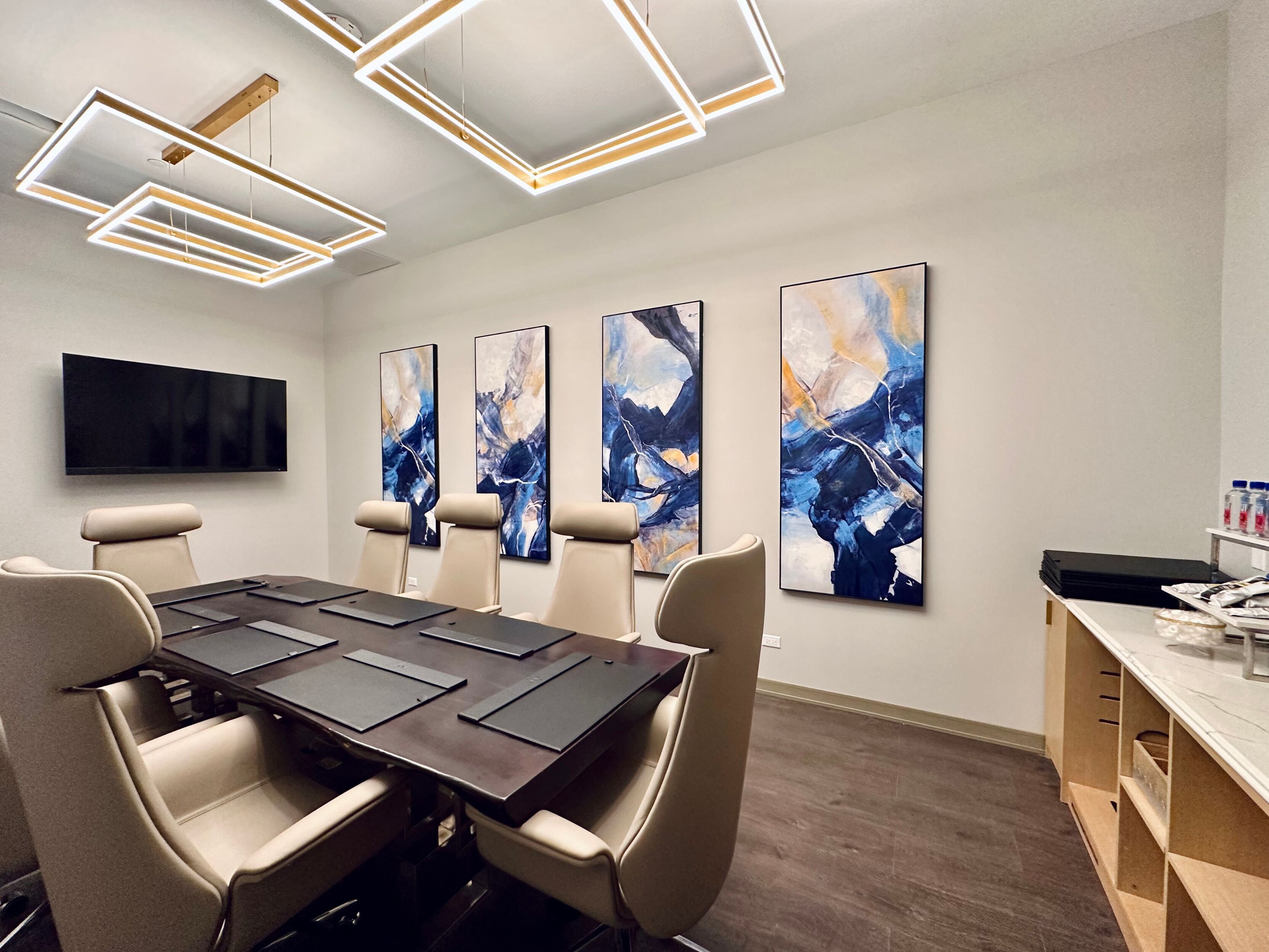 A conference room with a long table, four chairs, a flat-screen TV, and abstract paintings on the wall.