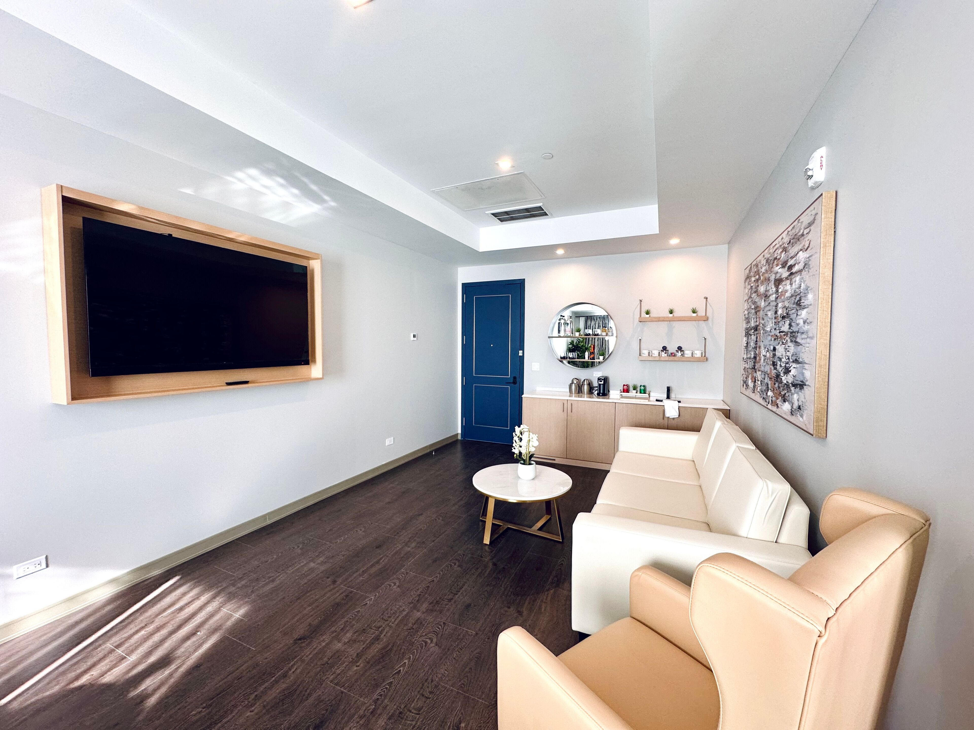 A modern living room with a large TV, a sofa, a coffee table, and a kitchen area with a blue door.