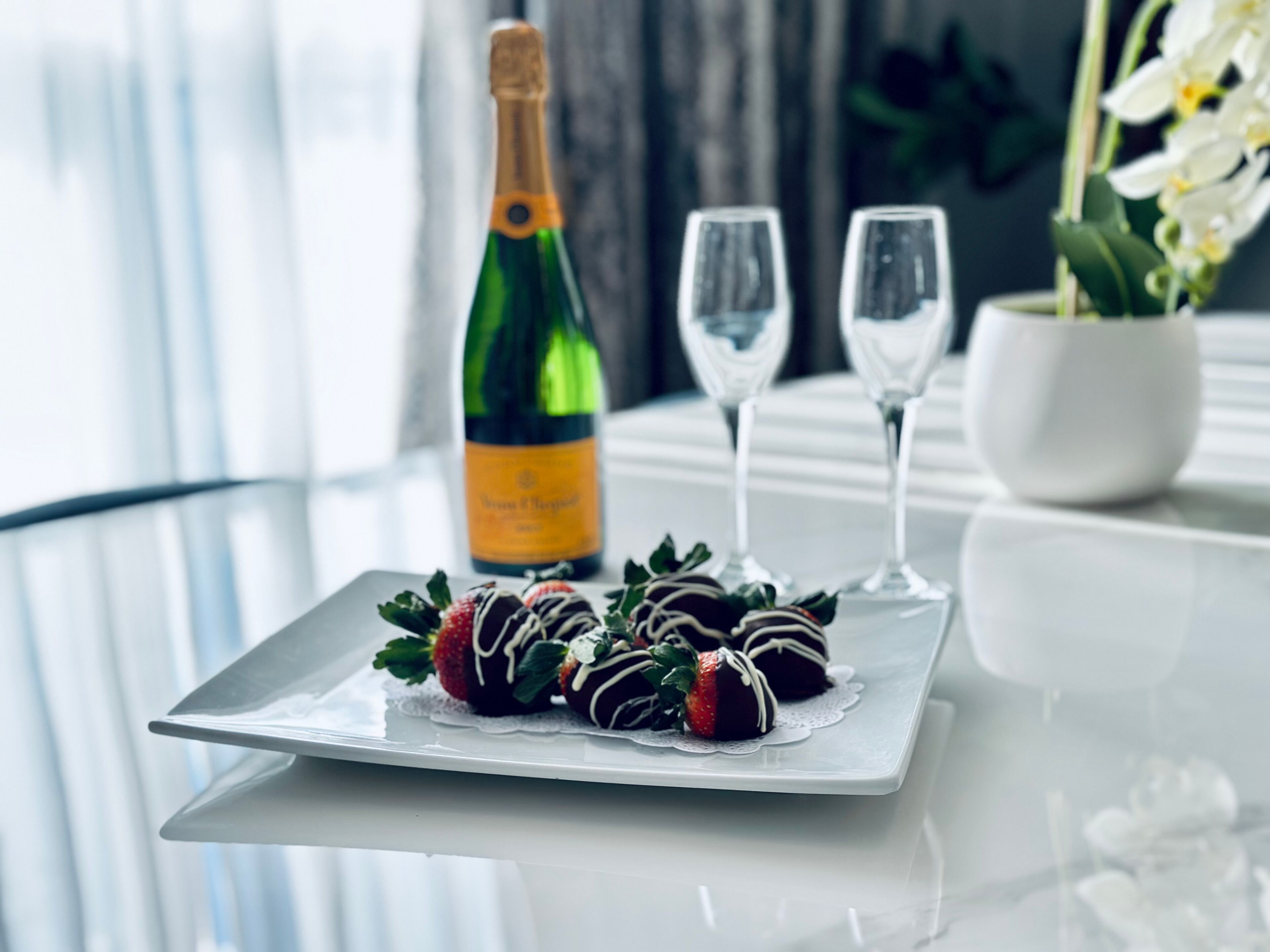 A glass table with a bottle of champagne, two glasses, and chocolate-covered strawberries on a white plate.