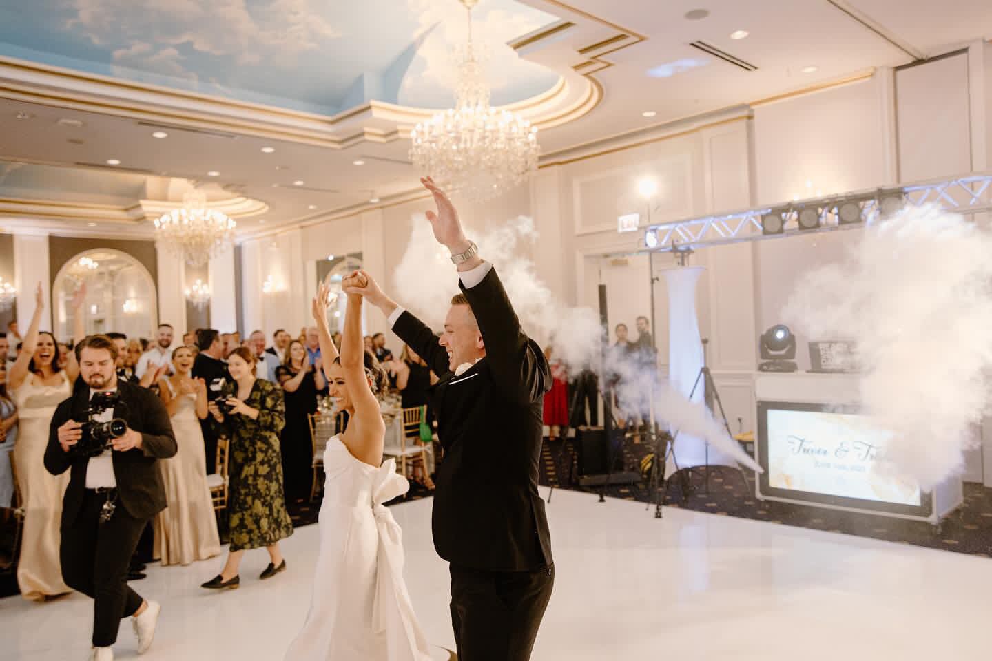 Eine Hochzeitsparty in einem geräumigen Saal mit einer Kristallleuchter, einer Bühne mit Raucherzeuger und einem Bildschirm, der das Hochzeitsthema zeigt.
