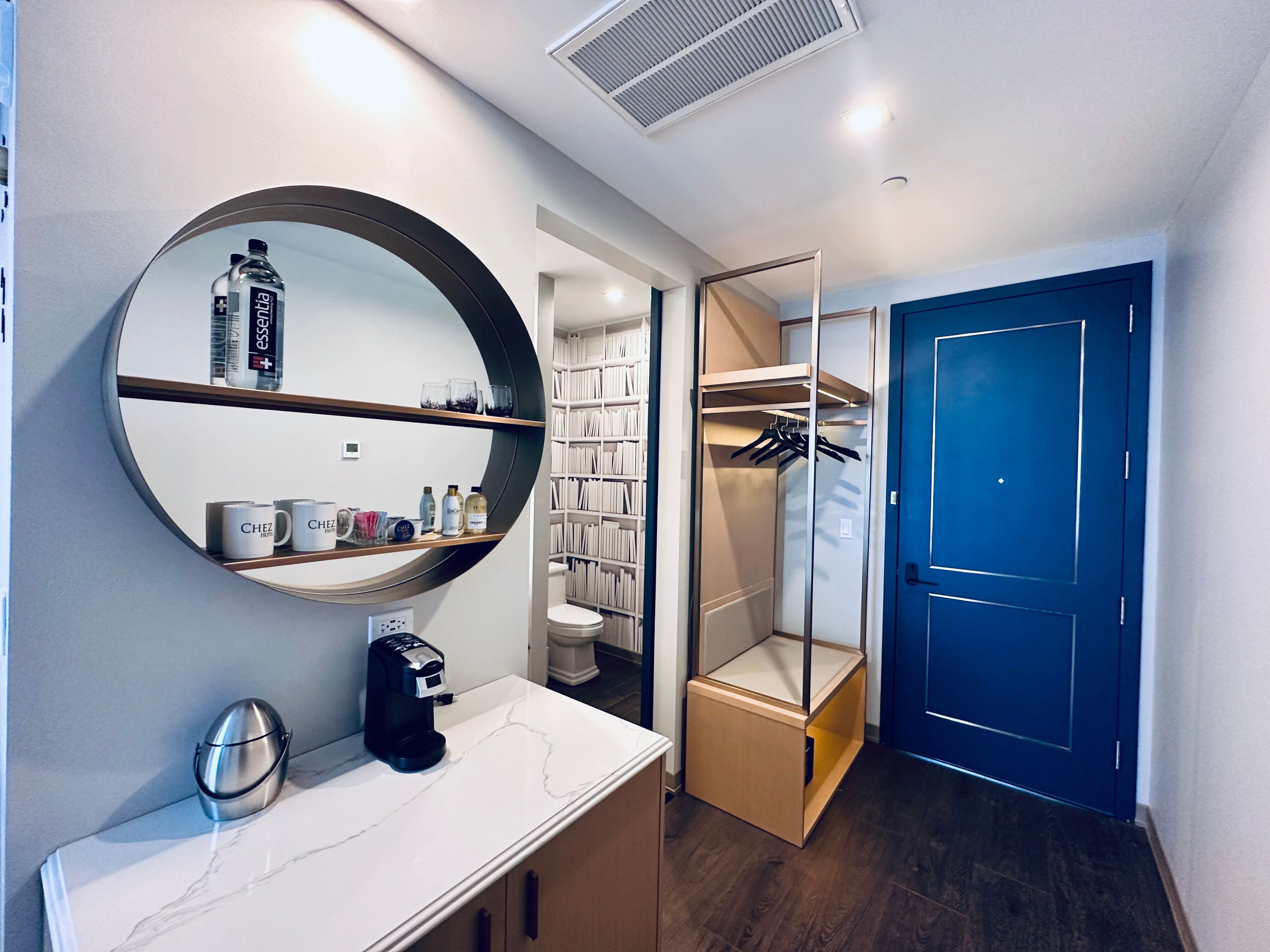 A modern bathroom with a round mirror, a coffee maker, and a shelf with toiletries.