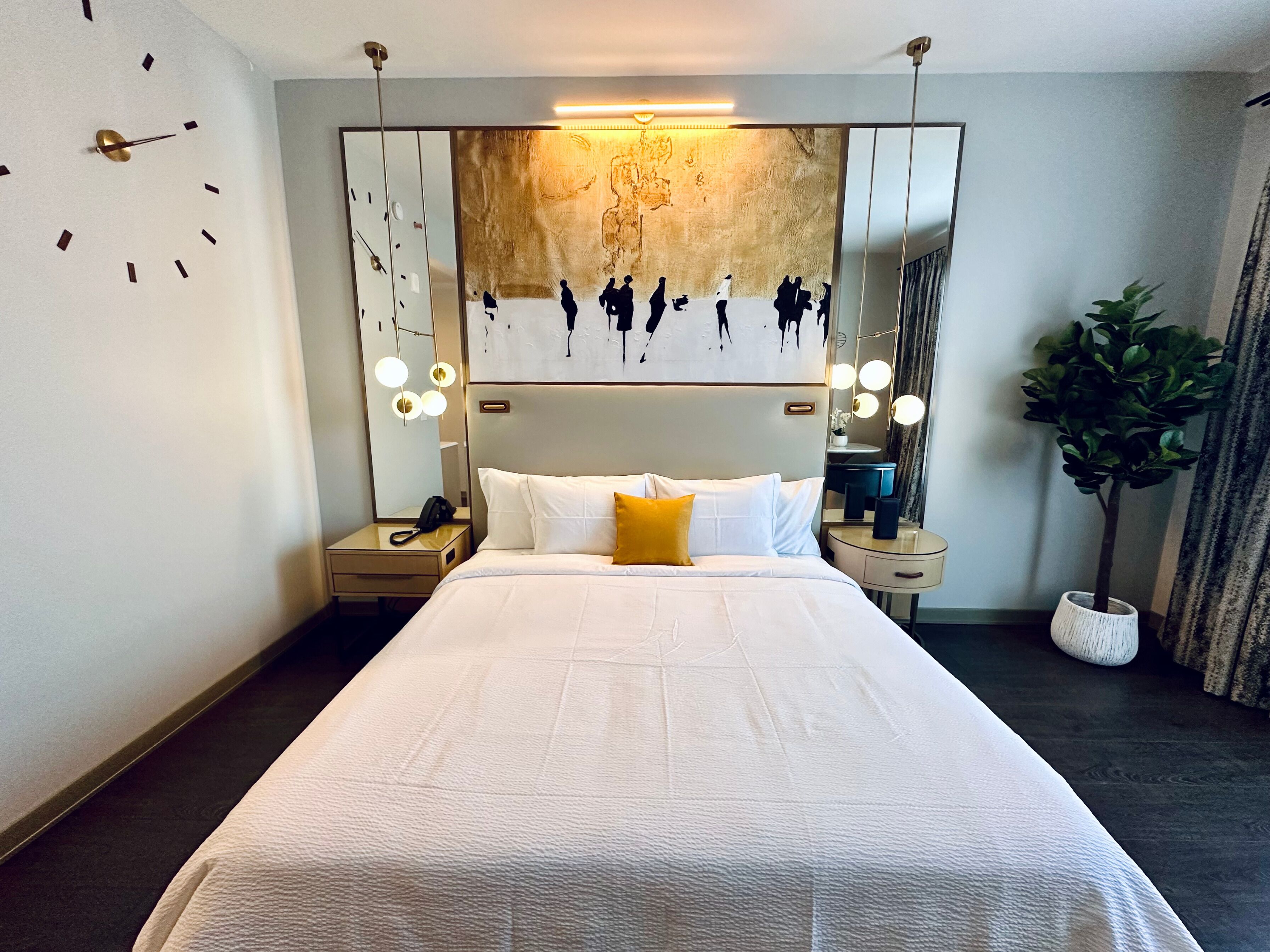 A modern hotel room with a large bed, a nightstand, a mirror with lights, and a wall clock.