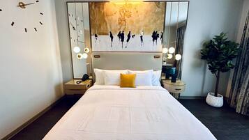 A modern hotel room with a large bed, a nightstand, a mirror with lights, and a wall clock.