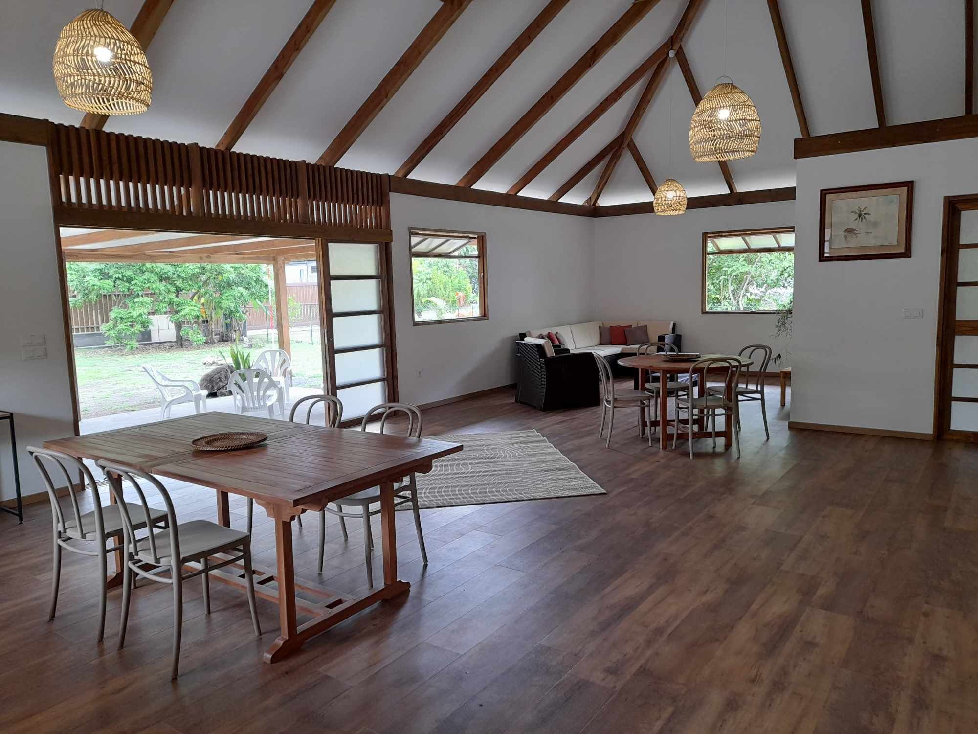A spacious room with a high ceiling, wooden beams, a dining area with a table and chairs, a sofa, and a view of the outdoors.