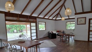 A spacious room with a high ceiling, wooden beams, a dining area with a table and chairs, a sofa, and a view of the outdoors. - Fare Pitate (Paea)