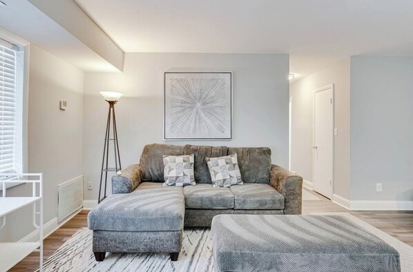 A modern living room with a grey sectional sofa, a matching ottoman, and a floor lamp. - 2BR Near Old Town & MGM First-Level Unit (Alexandria)