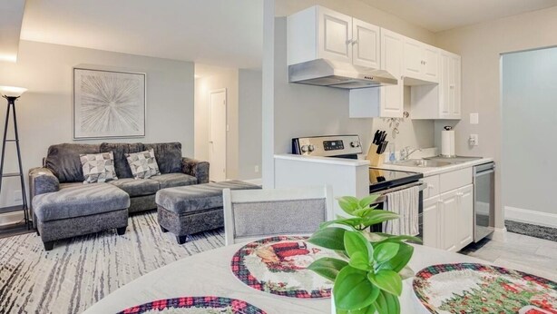 Un espacio habitable compacto con sofá, reposapiés y una mesa de comedor con individuales. Se observa una zona de cocina con armarios blancos y una estufa.