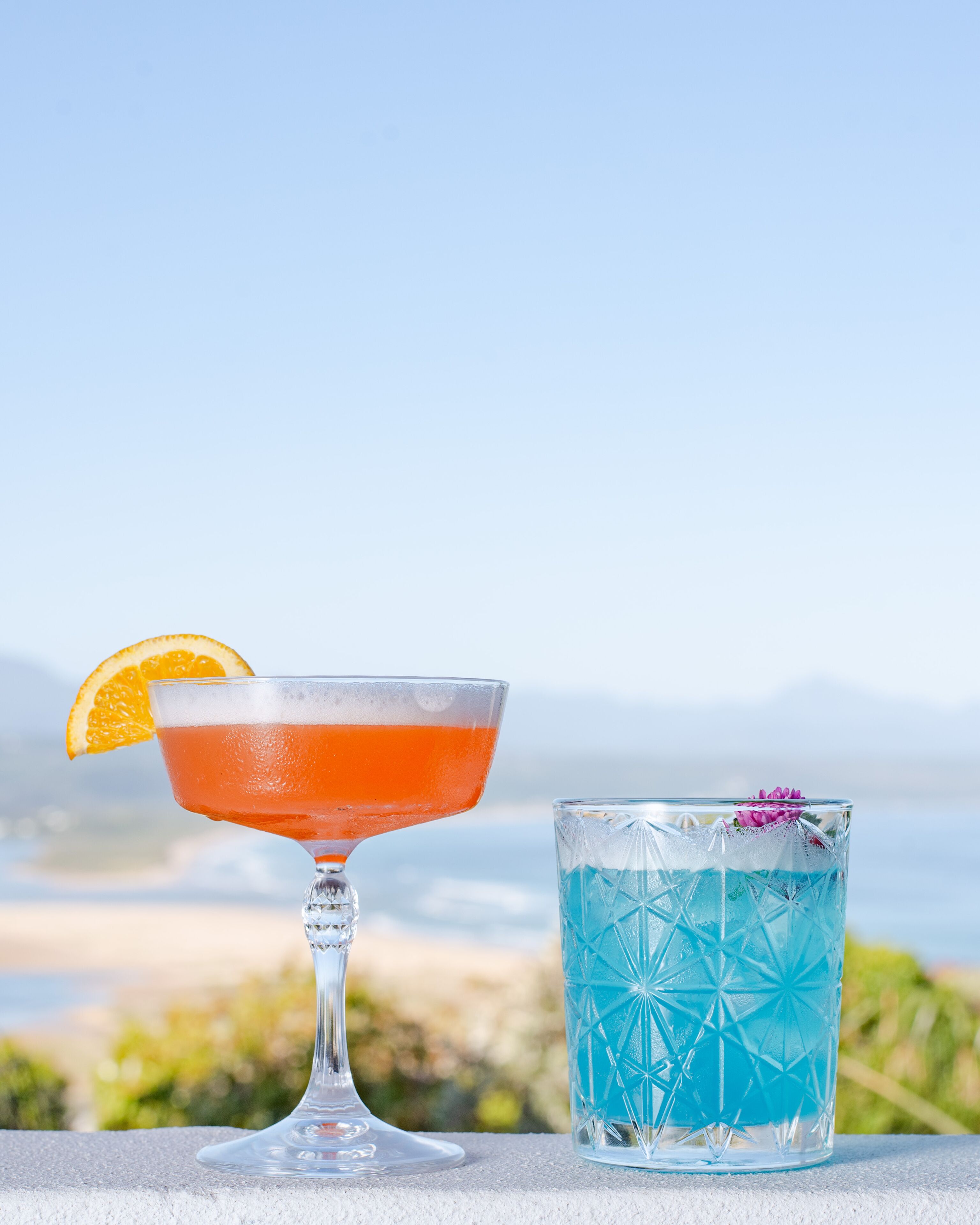 Two drinks on a table with a beach and mountains in the background.