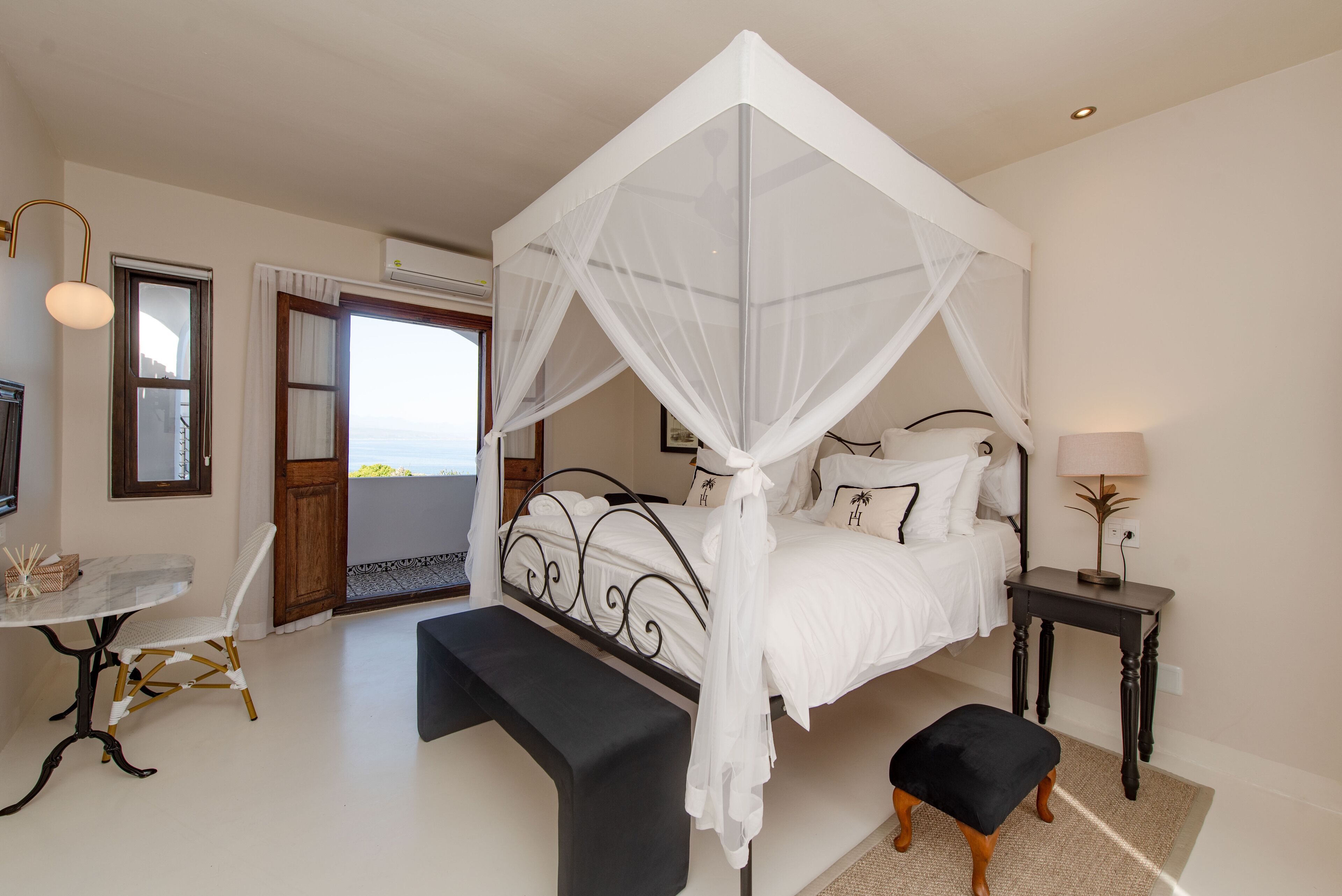A bedroom with a four-poster bed, a bedside table, a chair, and a view of the sea through a door.