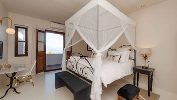 A bedroom with a four-poster bed, a bedside table, a chair, and a view of the sea through a door.