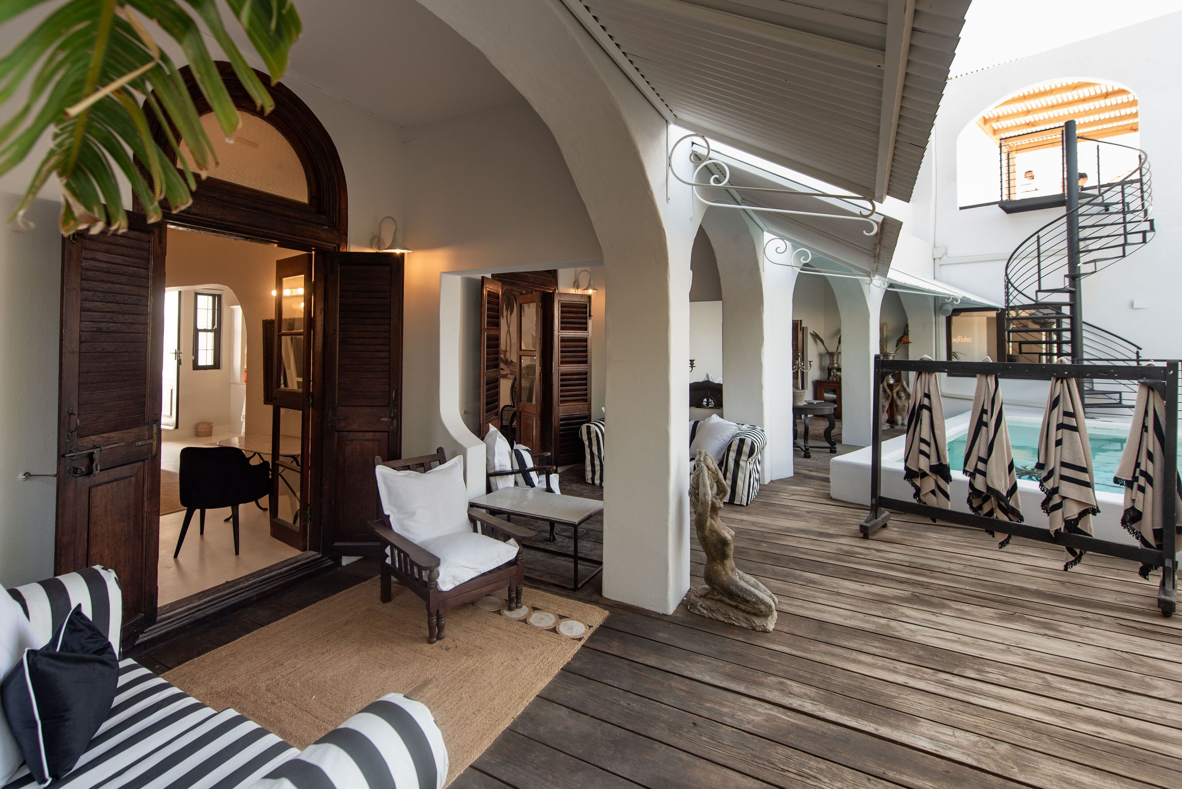 A spacious indoor area with a wooden deck, a seating area with a sofa and chairs, a staircase, and a pool area with lounge chairs.