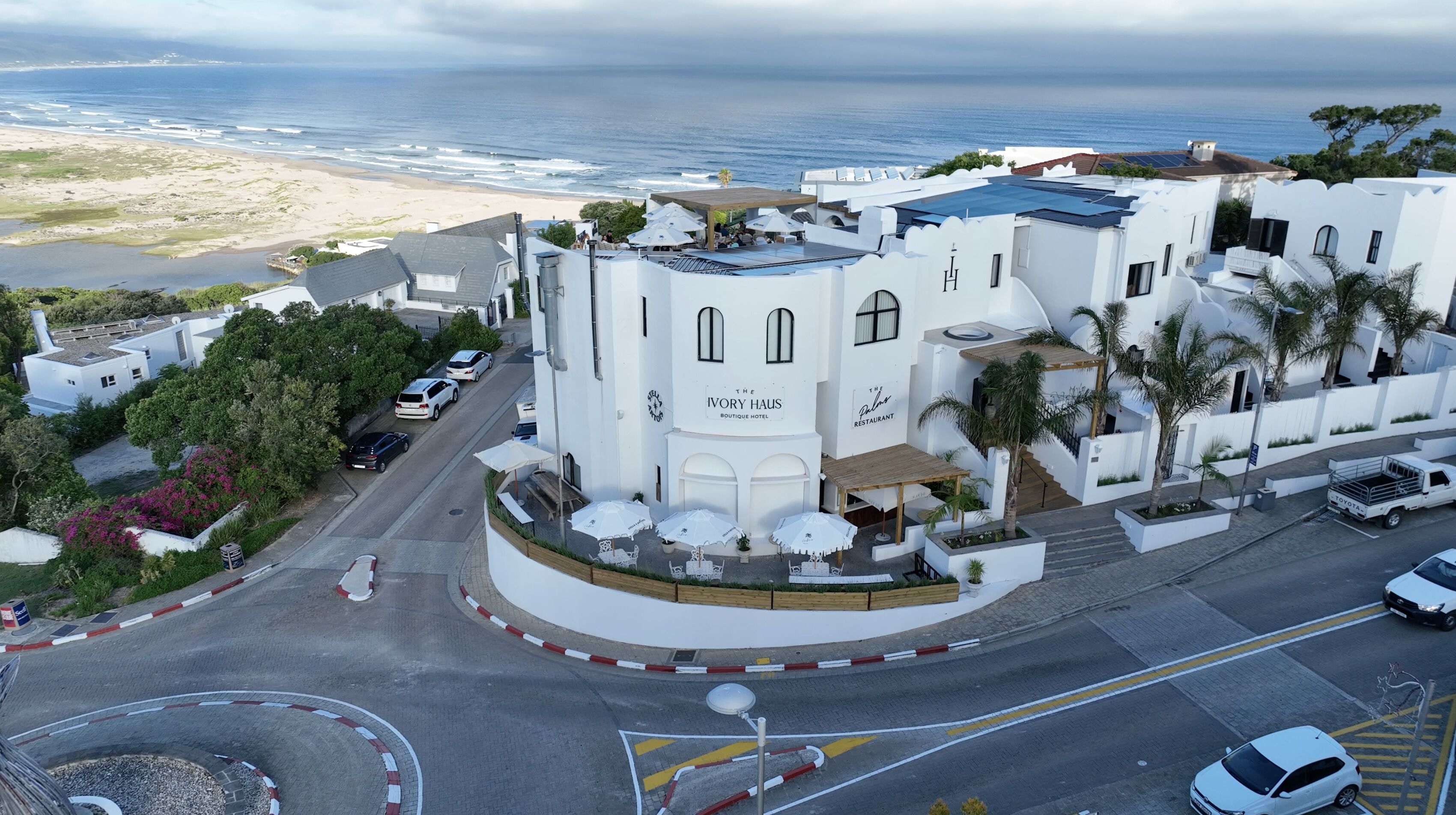 Un bâtiment blanc sur lequel on peut lire l’inscription « The Ivory Haus Boutique Hotel » et, sur le côté, « The Restaurant ». Il y a des espaces de restauration en plein air, avec des parasols, et une vue sur l’océan.
