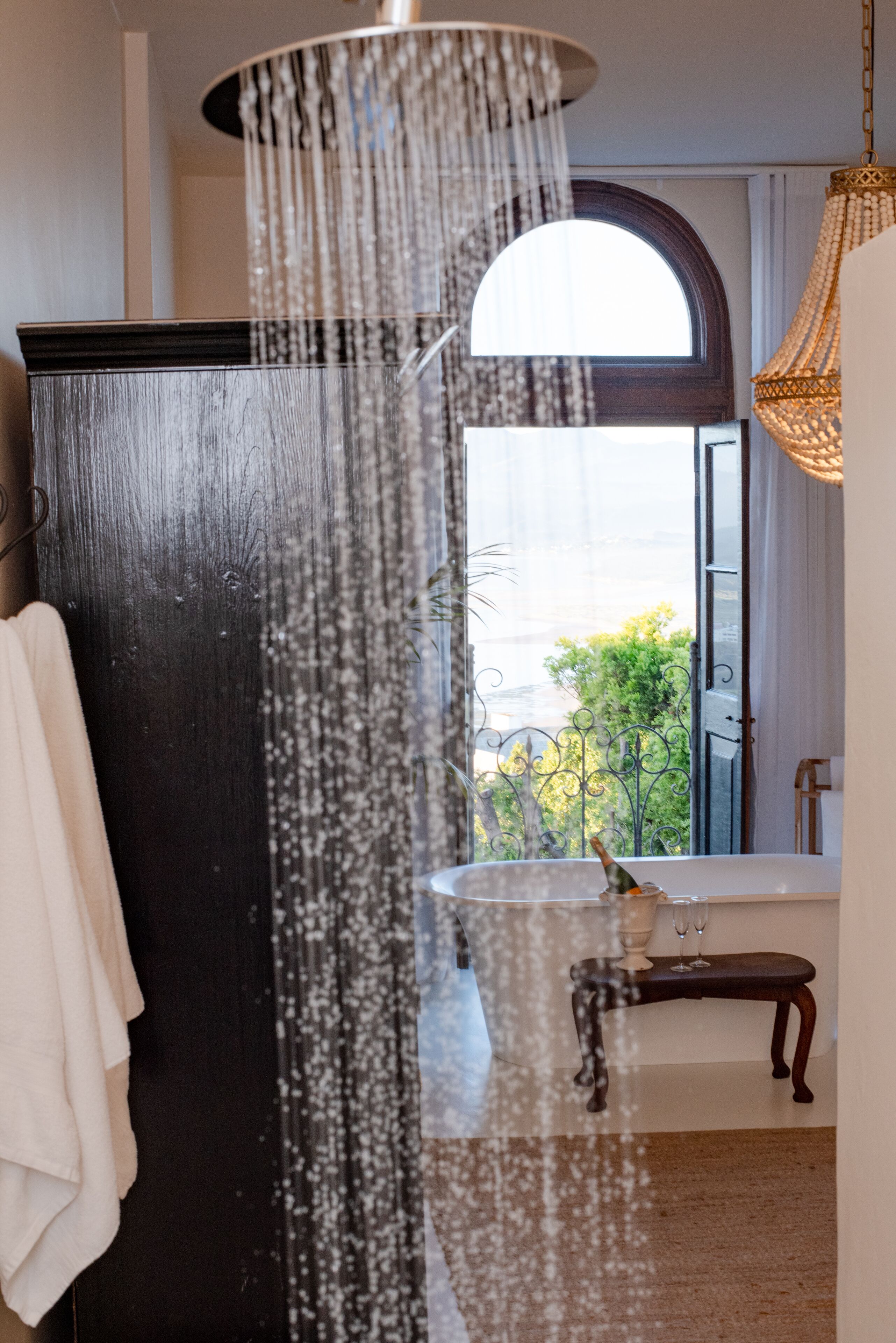 A bathroom with a bathtub, a shower with a waterfall feature, a window with a view of greenery, and a hanging light fixture.
