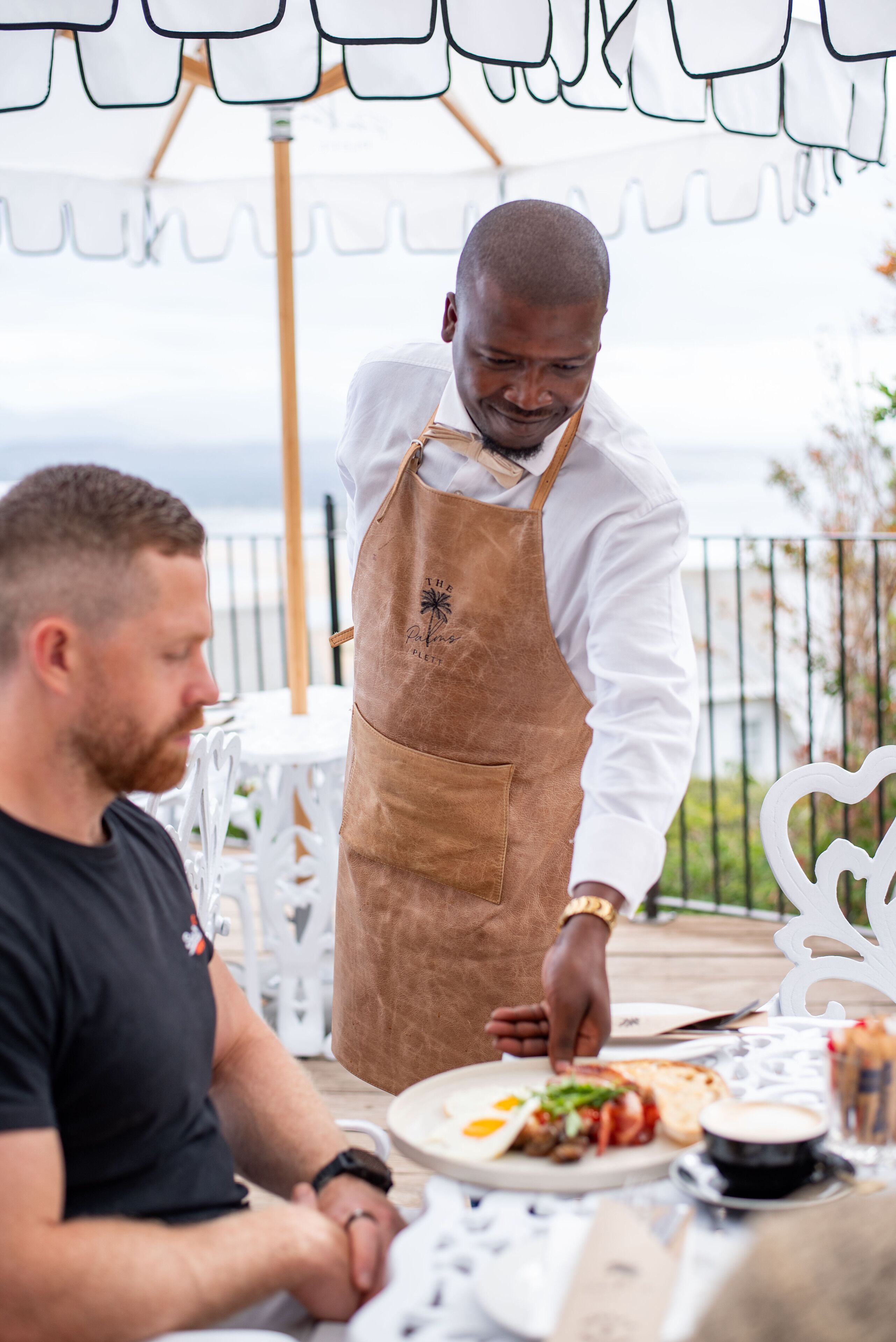 A person serving food at an outdoor table with a view of the sea.