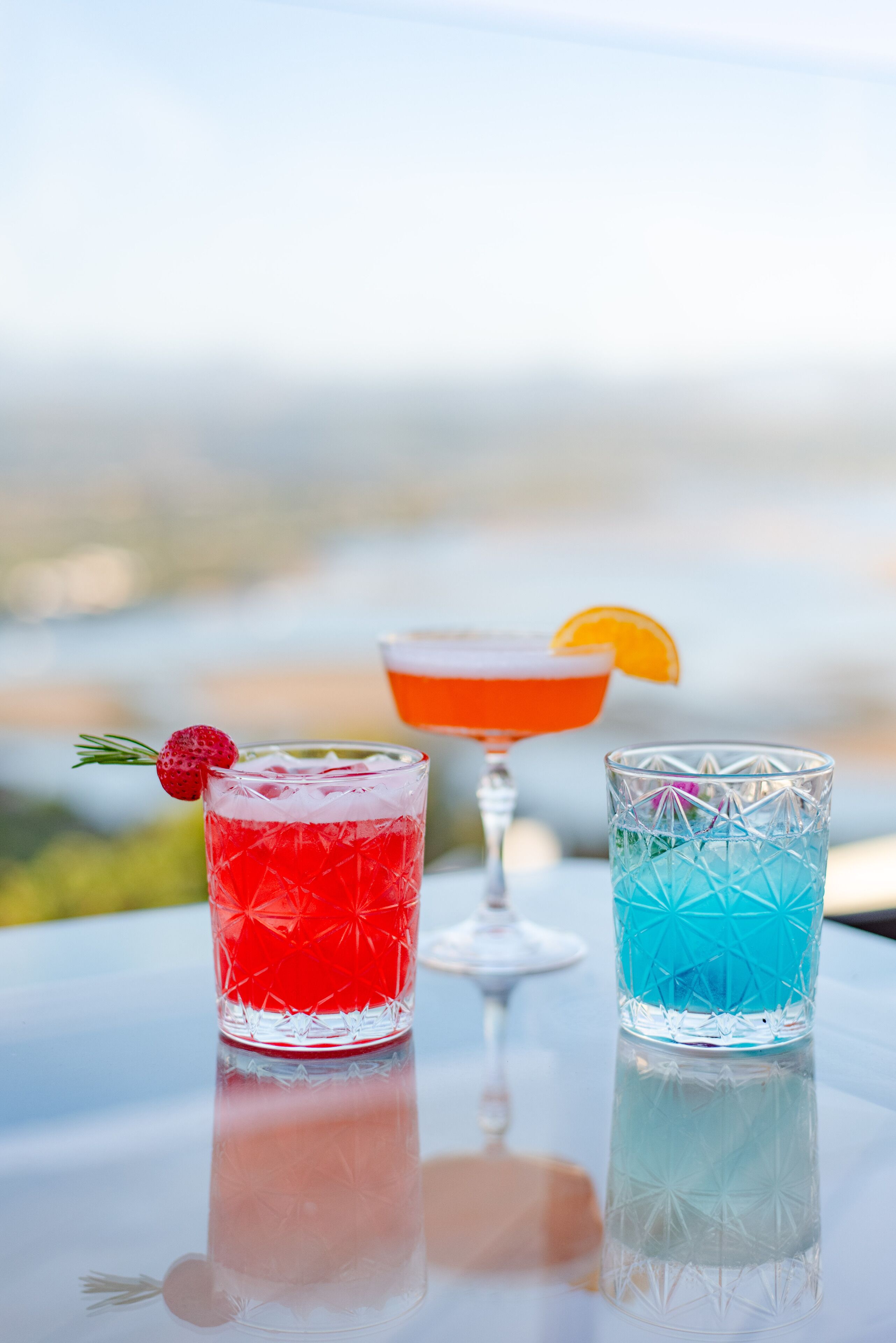 Three glasses of cocktails on a table with a view of the ocean.