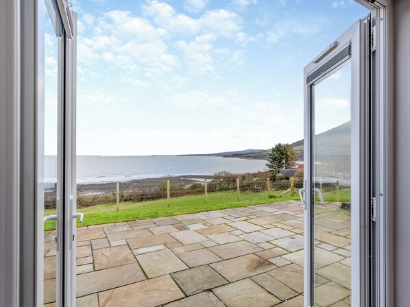 A view from a room with a glass door leading to a stone patio overlooking a body of water and grassy area.