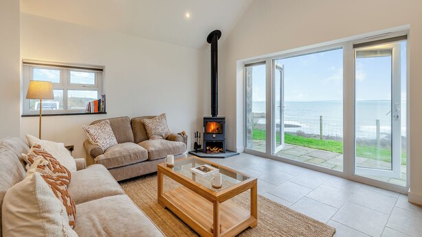 A cozy living room with a sofa, a coffee table, a fireplace, and a view of the sea through large glass doors. - 2 bedroom accommodation in Trefor Nr Caernarfon (Trefor Nr Caernarfon)
