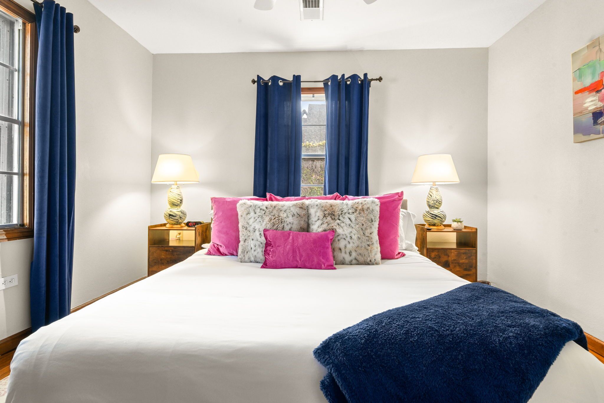 Ein ordentlich bezogenes Bett mit weißer und pinkfarbener Bettwäsche, einer blauen Tagesdecke und zwei Nachttischlampen.
