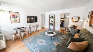 Un salon moderne avec un canapé, une table basse et un téléviseur. Il y a également un coin repas avec des chaises et une table. La pièce est agrémentée d’un grand tapis et d’une plante.