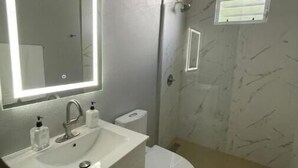 Une salle de bain avec un lavabo blanc, un miroir avec éclairage intégré, des toilettes et un espace douche.