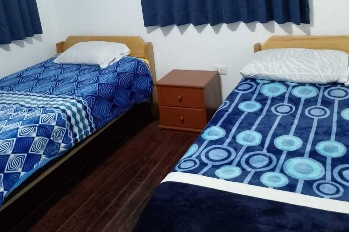 A room with two single beds, each with a patterned bedspread, a wooden nightstand, and a dark blue curtain covering a window.