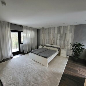 Une chambre à coucher moderne avec un grand lit, une table de chevet, une plante en pot et une vue sur l’extérieur par une fenêtre.