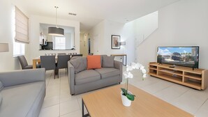 Una sala de estar moderna con un sofá gris, una mesa de centro con una planta, un televisor sobre un mueble de madera y una zona de comedor al fondo.