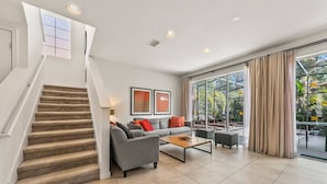 A spacious living room with a staircase, a grey sofa set, a coffee table, and large windows offering a view of the outdoors.