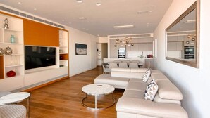 Un salon moderne avec une grande télévision, des canapés blancs, une table basse et un coin cuisine à l’arrière-plan.