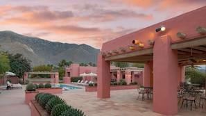 A pool area with lounge chairs and umbrellas, a covered seating area with tables and chairs, and a mountainous backdrop.