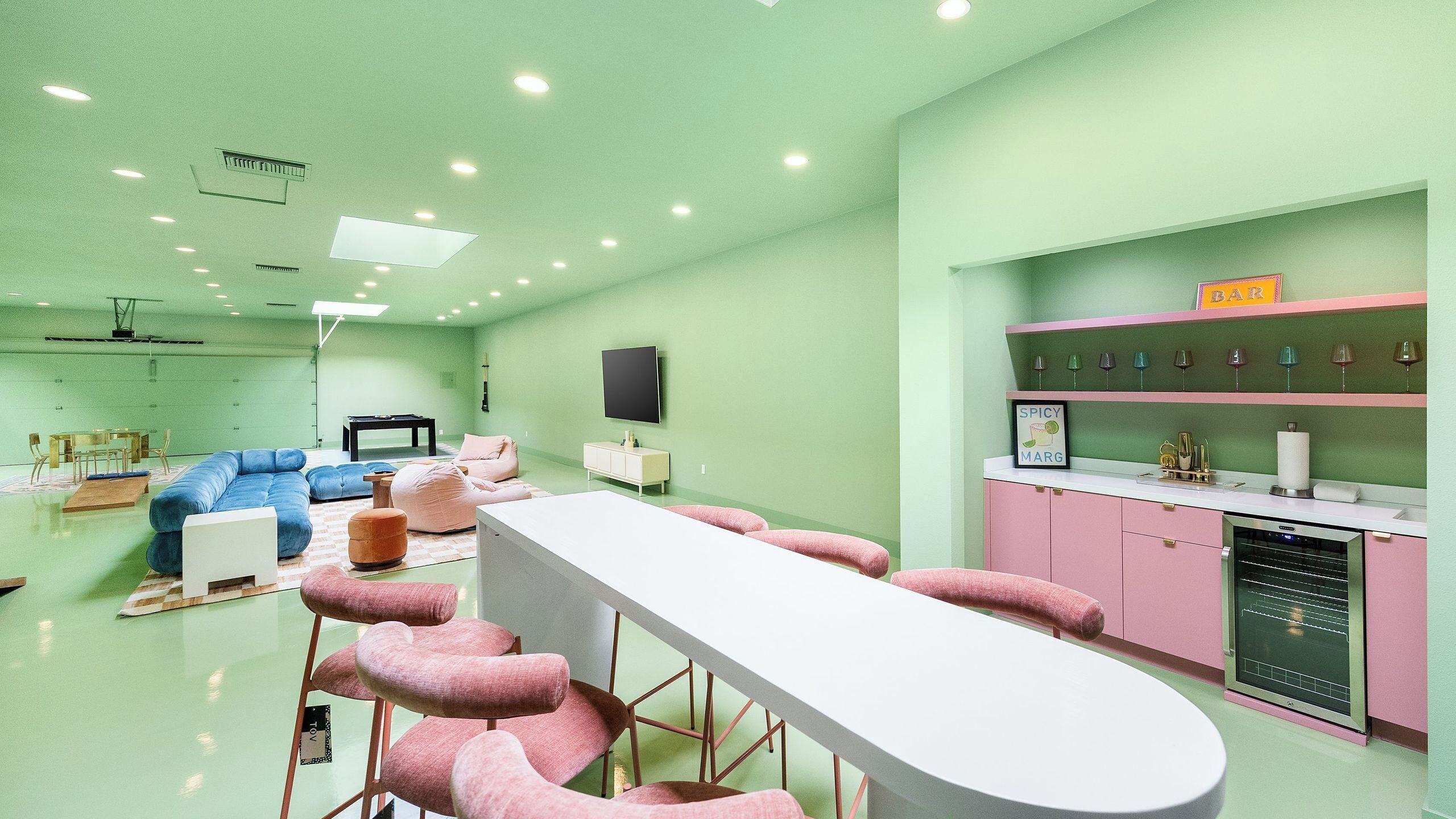 A room with a long white table, pink bar stools, a blue sofa, and a pink cabinet with wine glasses.